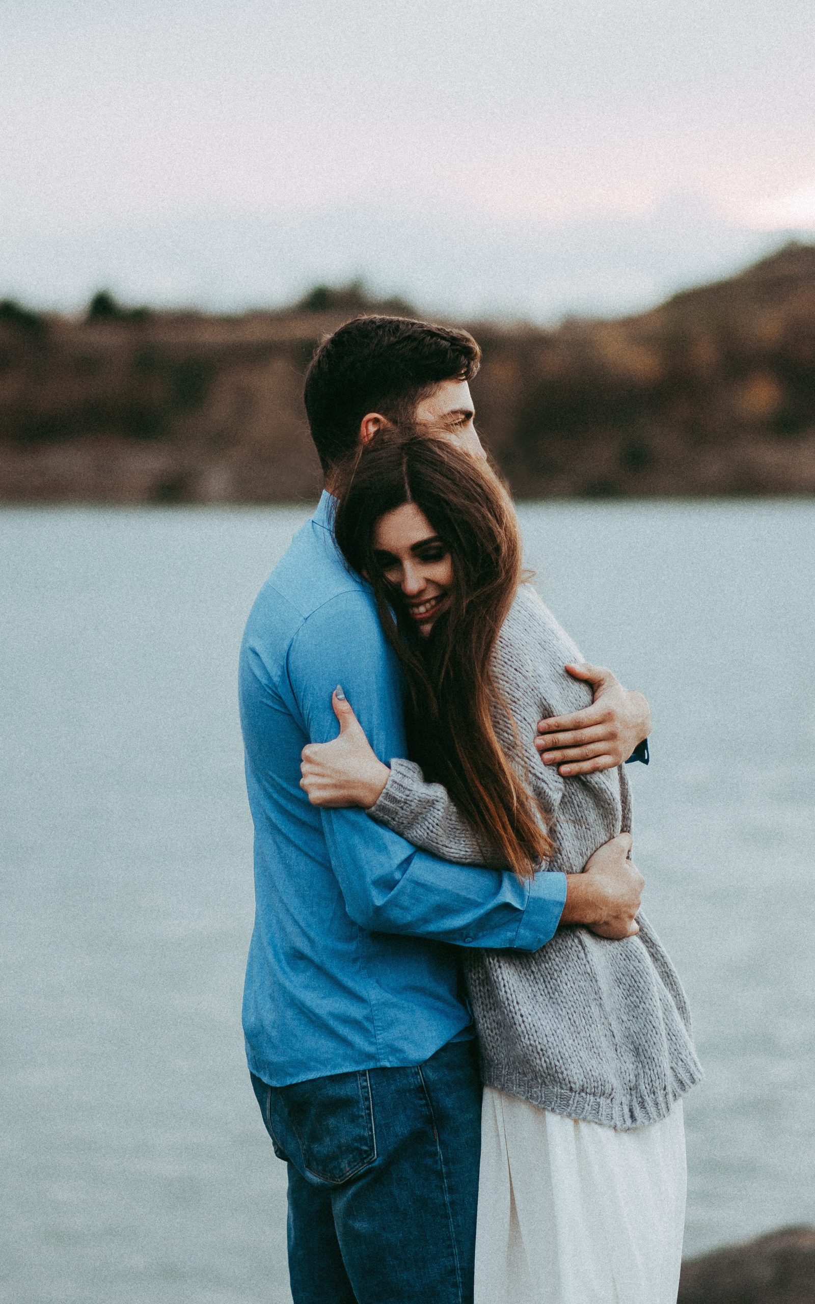 man-hugging-woman-wearing-gray-sweater-near-body-of-water-2071114